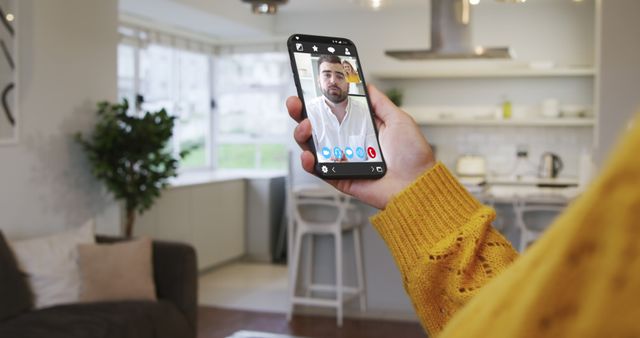 Person holding smartphone and making a video call with a man on the screen. Image is set in a bright, modern kitchen with a well-lit interior. Perfect for illustrating concepts of remote communication, modern technology, and staying connected with loved ones.