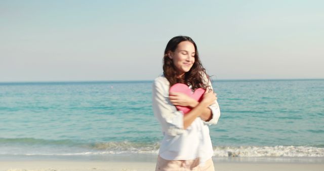 This image depicts a young woman embracing a heart-shaped pillow while standing on a serene beach. Ideal for themes of love, self-care, summer, and relaxation, with a calming ocean background that evokes warmth and happiness. Suitable for promoting wellness retreats, romantic getaways, self-love campaigns, or beach vacation packages.