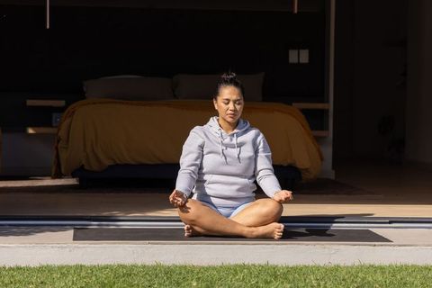 Asian woman meditating on home threshold wearing gray hoodie, morning sunlight, bedroom