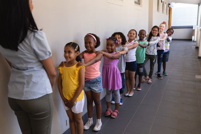 Schoolchildren standing in a line with their hands on each other's shoulders, led by a teacher in a school corridor. Ideal for educational materials, school brochures, and articles on childhood education, teamwork, and classroom discipline.