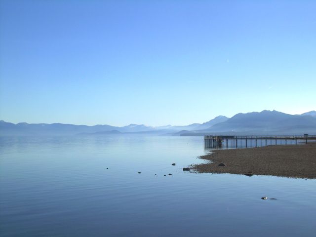 Image showcases a serene lakeshore setting with a wooden pier extending into the calm waters. Towering mountains are visible in the background under a clear blue sky. Perfect for travel blogs, nature websites, outdoor adventure promotions, and wellness retreats highlighting tranquility and natural beauty.