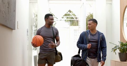 Young friends walking through bright foyer carrying basketball and gym duffel bags, chatting