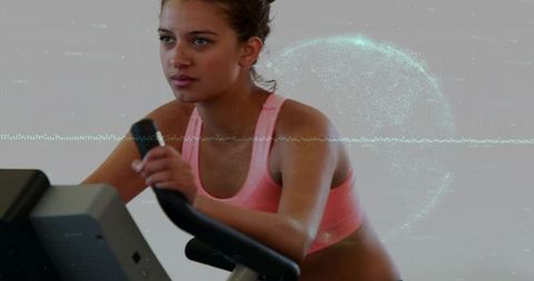 Woman cycling on stationary bike in pink sports bra with teal digital overlay, cardio focus