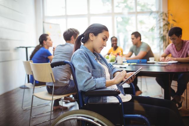 Businesswoman in a wheelchair using a digital tablet in a modern office environment. Colleagues in the background engaged in a meeting. Ideal for illustrating concepts of diversity, inclusion, technology in the workplace, and professional collaboration.