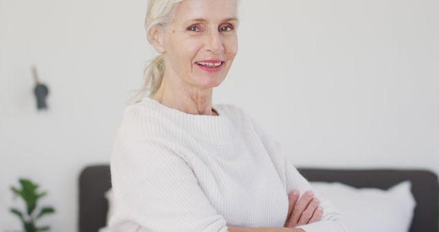 Senior woman with gray hair wearing a cozy sweater sits at home with arms crossed, displaying confidence and contentment. The bright indoor setting adds warmth to the calm, comfortable atmosphere. Ideal for use in lifestyle blogs, articles focused on aging gracefully, and home living advertisements.