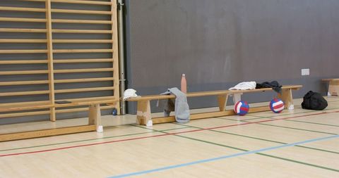 Wooden bench lining gym wall holding workout clothes and two volleyballs on indoor court