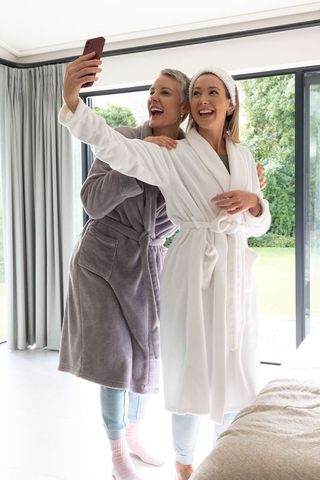 Mother and daughter taking selfie in sunlit bedroom wearing plush bathrobes