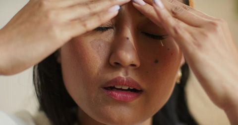 Asian woman pressing fingertips to forehead, shedding tear, reflecting on stress