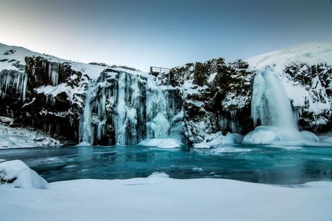 Icy waterfall cascading into turquoise frozen pool amid snow-covered cliffs and icicles