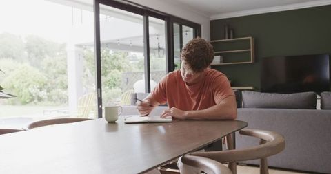 Young man writing journal at dining table by glass doors in bright modern living room