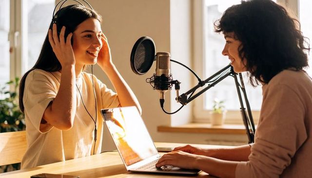 Co-hosts recording podcast in bright home studio adjusting headphones and using laptop