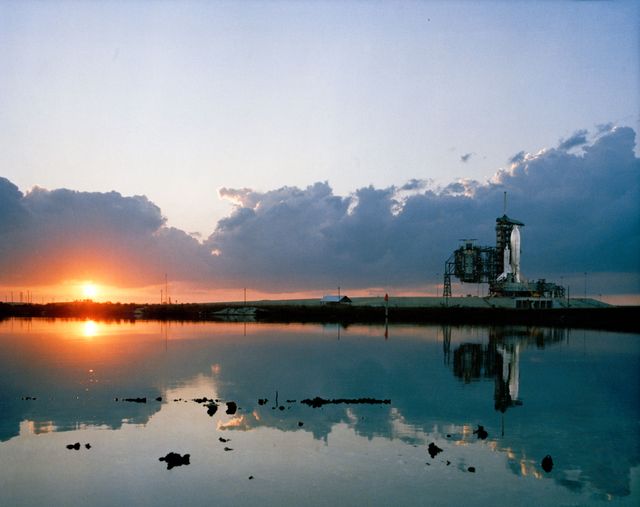 Space shuttle Columbia is positioned on Launch Pad A at Kennedy Space Center during sunrise in March 1981. The peaceful early morning scene reflects Columbia's image in a body of water, capturing the anticipation before the historic STS-1 mission. This image is suitable for educational materials, aerospace industry reports, and articles about space exploration history.