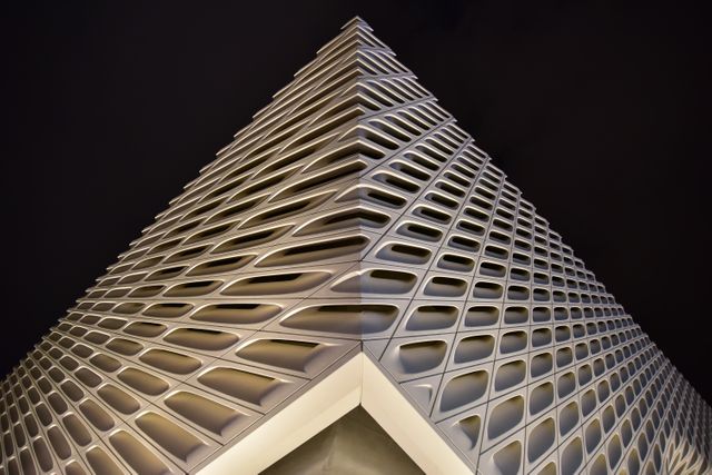 The image shows the corner of a modern architectural structure with a unique geometric pattern at night, with lit windows and a dark sky. The dramatic angle emphasizes the design details. Ideal for use in articles about architectural design, urban planning, construction, and contemporary architecture features.