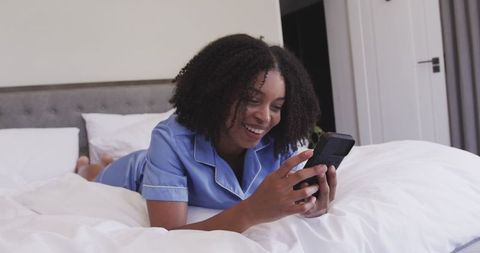 Smiling woman in blue pajamas lying on bed scrolling smartphone in cozy bedroom
