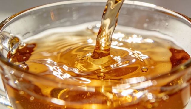 Golden amber liquid pouring into glass cup closeup showing ripples, bubbles and reflection