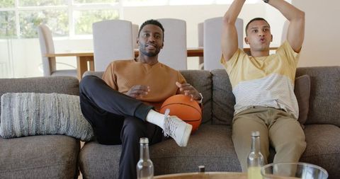 Diverse friends watching basketball at home on sofa holding ball cheering casual weekend