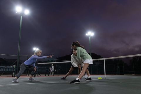Female tennis teammates practicing drills at dusk under floodlights on outdoor hard court
