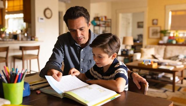 Father guiding son through homework at kitchen table, tutoring and reading together