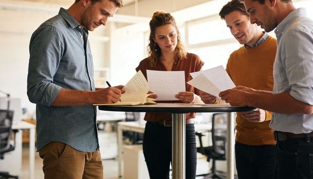 Startup team reviewing documents and taking notes around standing round table in open-plan office
