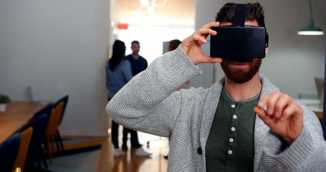 Visual depicts a bearded man in casual clothing using a virtual reality headset in modern office space. Great for illustrating technology use in professional environments, digital workplace concepts, VR innovation, and trends in interactive media. Useful for tech blogs, articles on workplace innovation, or promotional content for tech companies integrating VR.