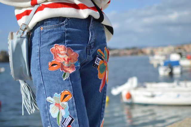 Close-up captures vibrant and fashionable patched jeans worn by a woman with a striped sweater by a waterfront. Suitable for fashion blogs, advertising campaigns for clothing brands, or editorial pieces on summer fashion and casual style.