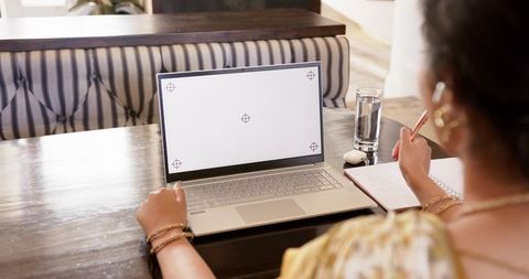 Indian woman in sari working remotely on laptop in cafe taking notes with earbuds