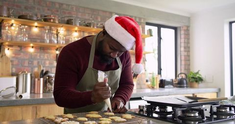 African American man wearing Santa hat piping icing on Christmas cookies in cozy kitchen