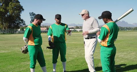 Senior coach coaching diverse baseball team in green uniforms on sunny community field