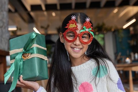 Preteen asian girl wearing red glitter christmas tree glasses smiling holding green gift