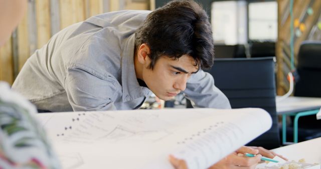 A young architect is intently examining a blueprint in a modern office environment. The setting is conducive for detailed design work, indicated by the presence of professional drafts. Ideal for articles, advertisements, and posts related to architecture, engineering, youthful workforce, and professional diligence.