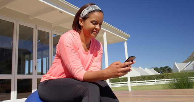 Woman sitting outdoors on a patio, dressed casually in athletic wear, engrossed in her smartphone. Ideal for themes related to relaxation, technology, leisure activities, modern lifestyle, outdoor living, and mobile connectivity.