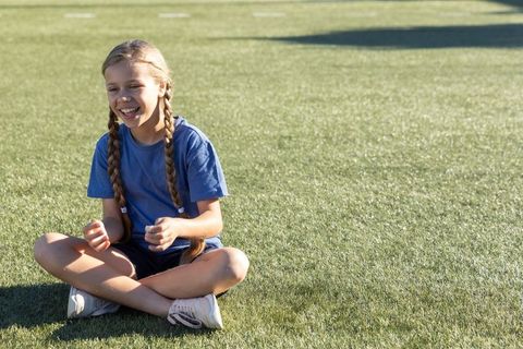 Smiling girl with braids sitting cross-legged on turf wearing blue tee and white sneakers