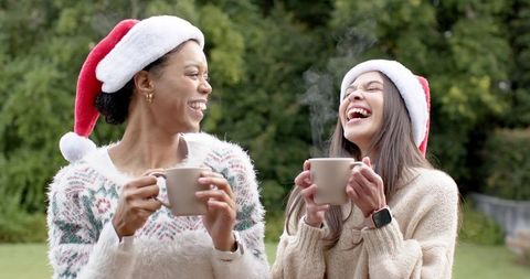 Friends laughing with steaming mugs in garden wearing santa hats and cozy knit sweaters