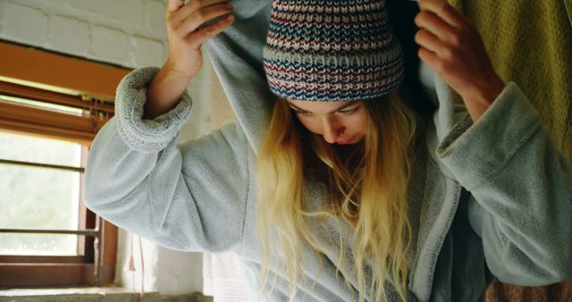 A woman is putting on a warm hoodie while wearing a beanie. This image captures a cozy indoor moment, suggesting a cold weather comfort strategy. This image can be used for articles or advertisements centered around winter clothing, home comforts, loungewear, and morning routines.