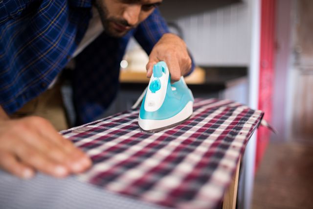 Man ironing a plaid shirt on an ironing board at home. Ideal for illustrating household chores, domestic tasks, and home life. Useful for articles, blogs, and advertisements related to laundry, home care, and lifestyle.