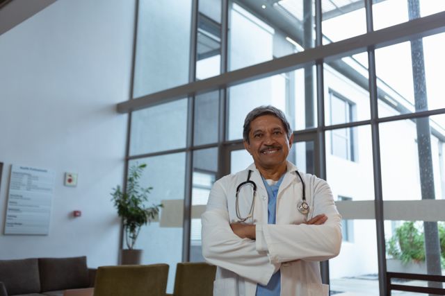Front view of happy mature male doctor with arms crossed standing in lobby at hospital