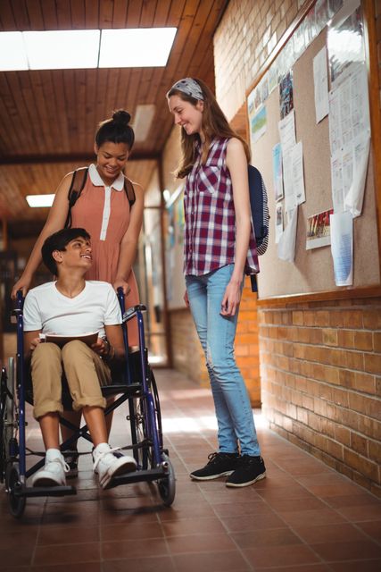 Two schoolgirls are talking with their disabled friend who is in a wheelchair in a school corridor. They are smiling and appear to be having a friendly conversation. This image can be used to promote themes of friendship, inclusion, diversity, and accessibility in educational settings. It is suitable for use in educational materials, social campaigns, and articles about inclusive education.