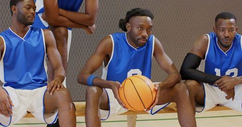 Sitting basketball teammates holding orange ball on bench in gym wearing blue jerseys
