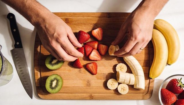 Hands slicing banana and arranging strawberries on wooden cutting board for smoothie prep