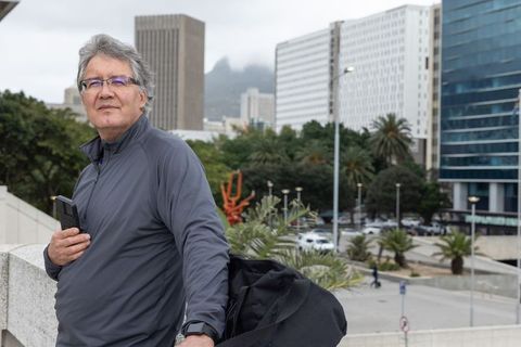 Senior man leaning on railing at city plaza holding smartphone and duffel bag