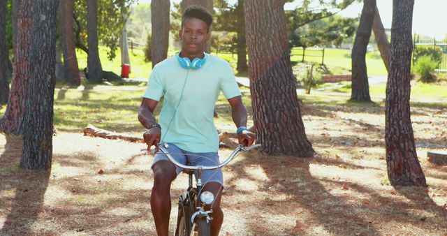 Young man riding bicycle through forest park surrounded by trees. Wearing headphones and casual clothing. Ideal for lifestyle, relaxation, outdoor activities, and nature themes.