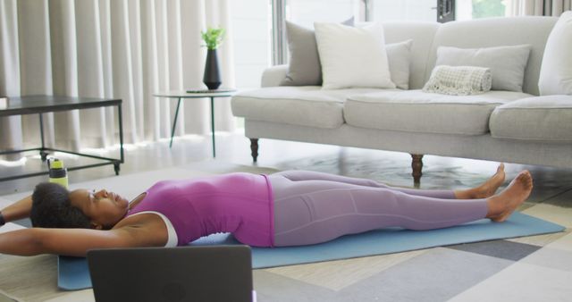 Woman practicing yoga on a blue mat in a modern living room. She is lying on her back with arms extended. The living room features a gray sofa and a coffee table. Ideal for use in fitness blogs, wellness websites, home workout inspiration, and lifestyle magazines.