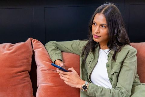 Young woman sitting on rust velvet sofa holding smartphone and contemplating in lounge