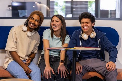 Laughing diverse students collaborating in modern study lounge with notebooks, headphones