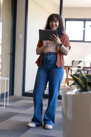 Smiling woman holding tablet standing in sunlit coworking lounge, casual productivity