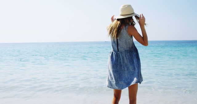 A young Caucasian woman in a summer dress and hat stands facing the ocean, with copy space. Her relaxed posture and the serene beach setting evoke a sense of calm and leisure.