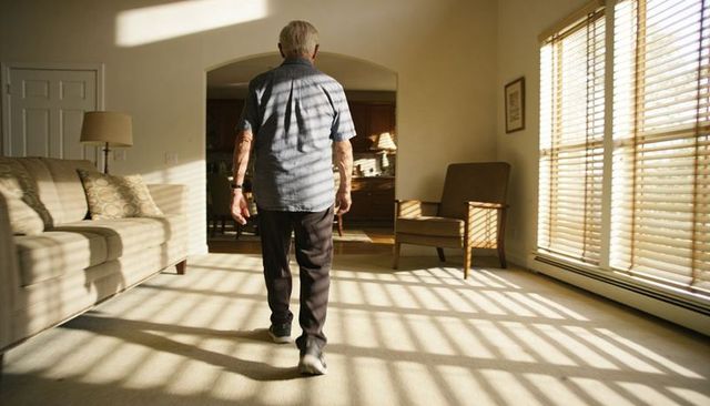 Senior man walking through sunlit living room with wooden blinds casting striped shadows