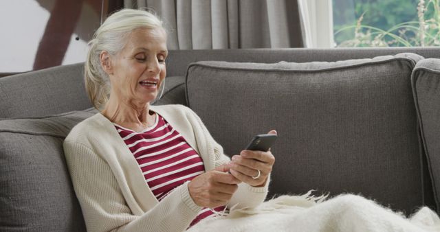 Senior woman relaxing on a couch while using a smartphone. The woman is wearing a cardigan over a red and white striped shirt and looks comfortable at home. Ideal for use in blogs, websites, and articles about senior lifestyle, technology adoption by seniors, home comfort, and retired life.