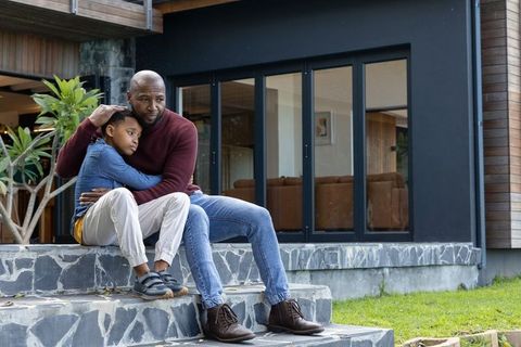African american father comforting son sitting on stone steps outside modern home