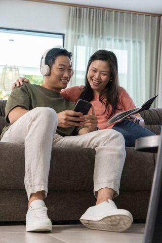 Asian couple relaxing on sofa sharing smartphone and laughing in modern living room